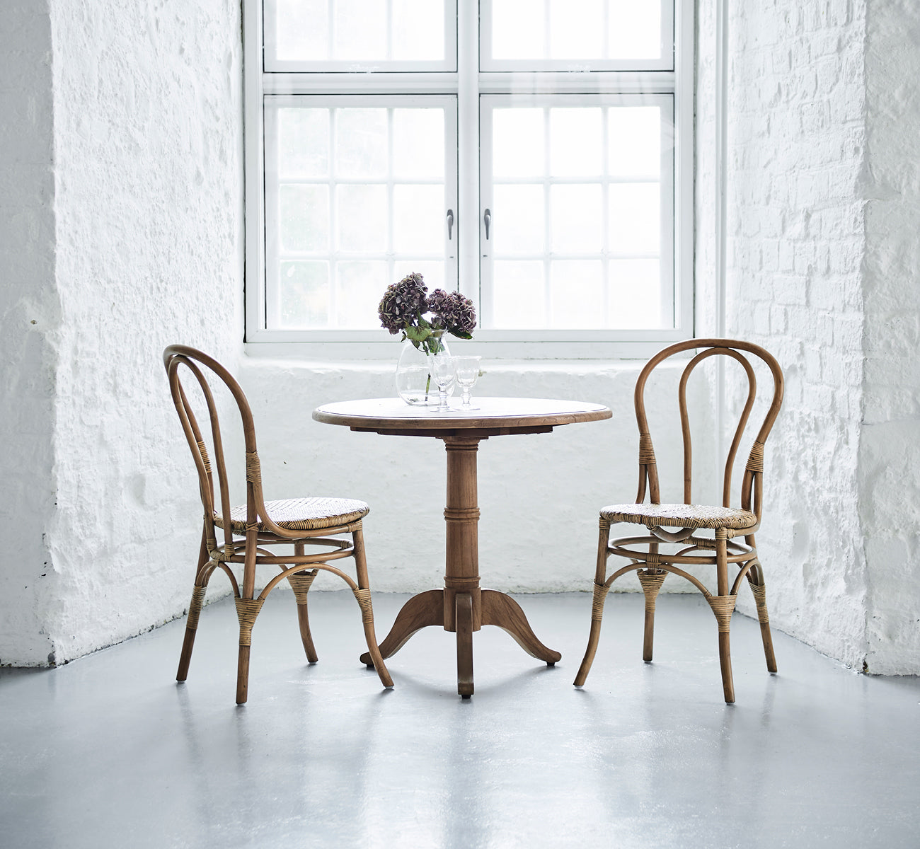 Dette er et livsstilsbillede af Lulu Spisebordsstol i antique rattan. To stole flankerer et rundt bord med blomster i et lyst hvidt murstensinteriør.