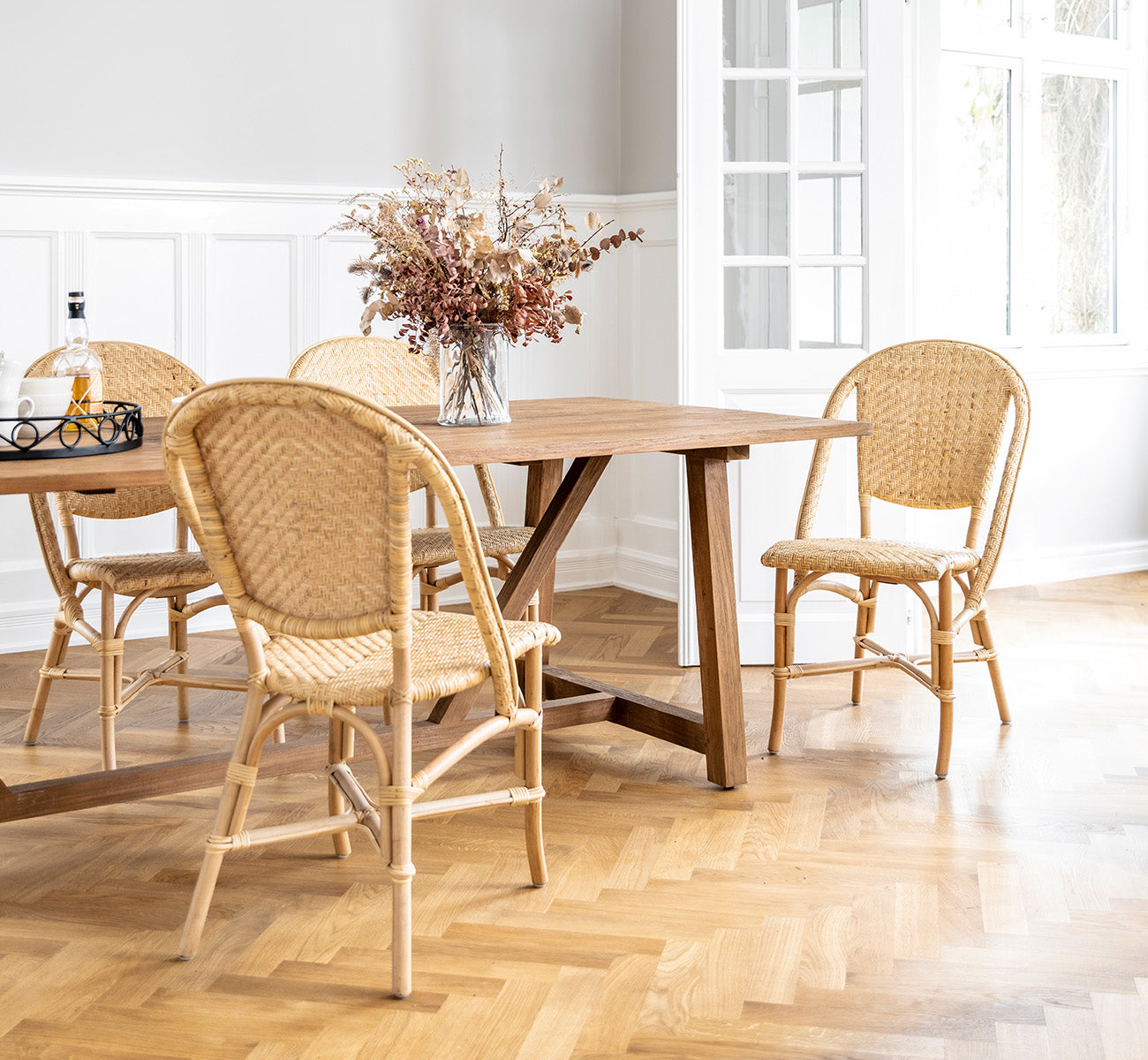 Dette er et livsstilsbillede af Alanis Spisebordsstol i natural rattan, i et lyst interiør med hvide panelvægge og sildebensgulv.