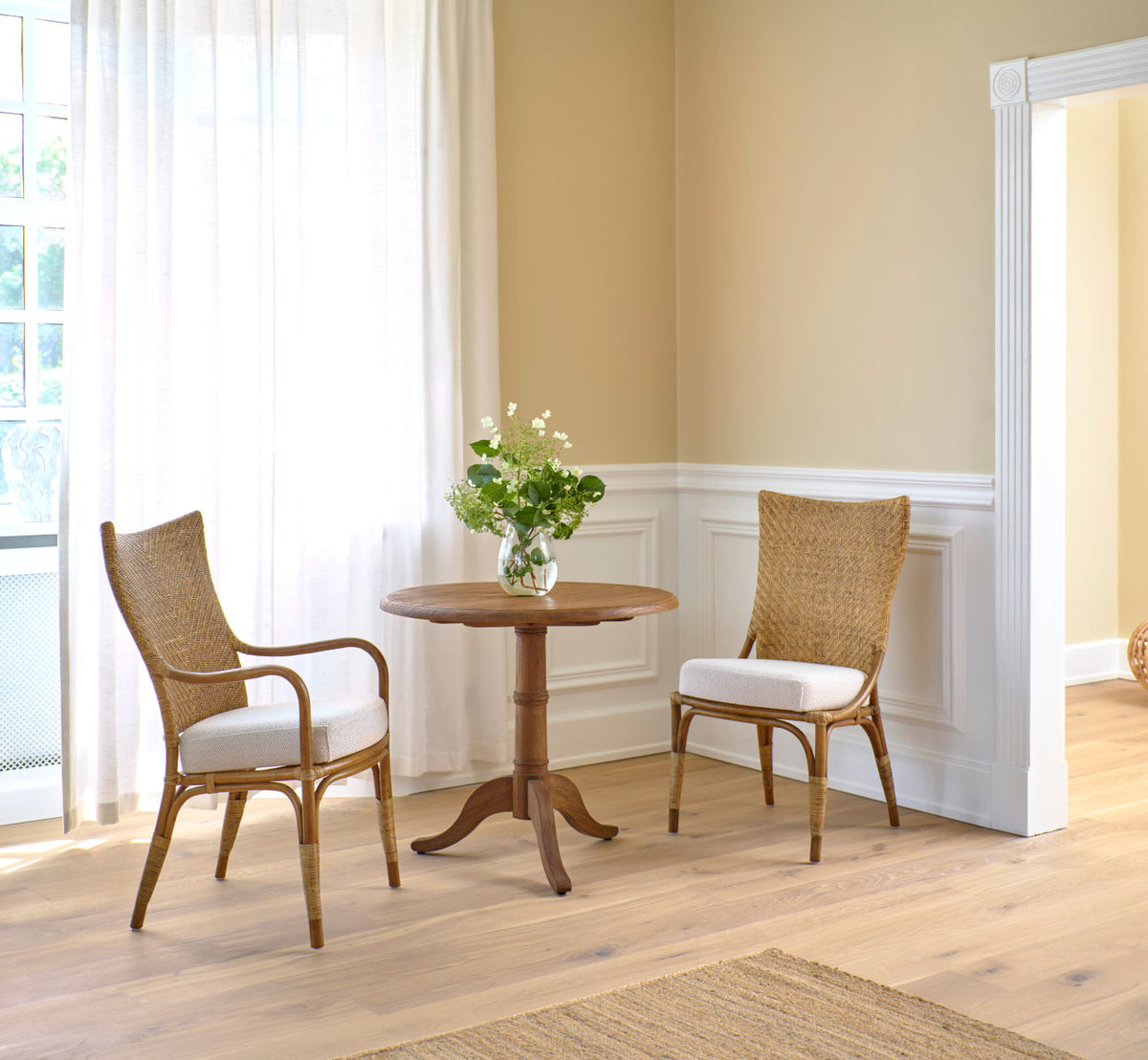 Dette er et livsstilsbillede af Melody Spisebordsstol i antique rattan. To ved et teak cafébord ved hvide paneler.