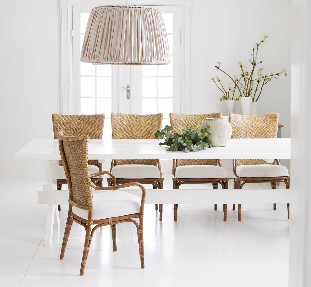 Dette er et livsstilsbillede af Melody Dining Armchair i antique rattan. Omkring et hvidt bord med linnedhængelampe og forårsgrene.