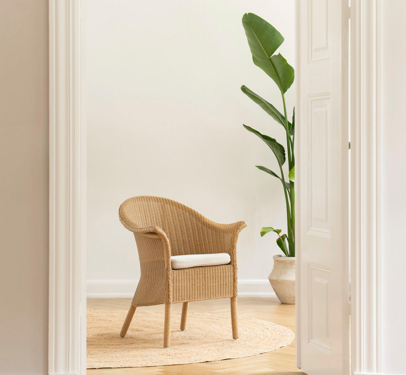 Dette er et livsstilsbillede af Classic Spisebordsstol i natural Loom Rattan Frame, i et lyst interiør med hvide panelvægge og sildebensgulv.
