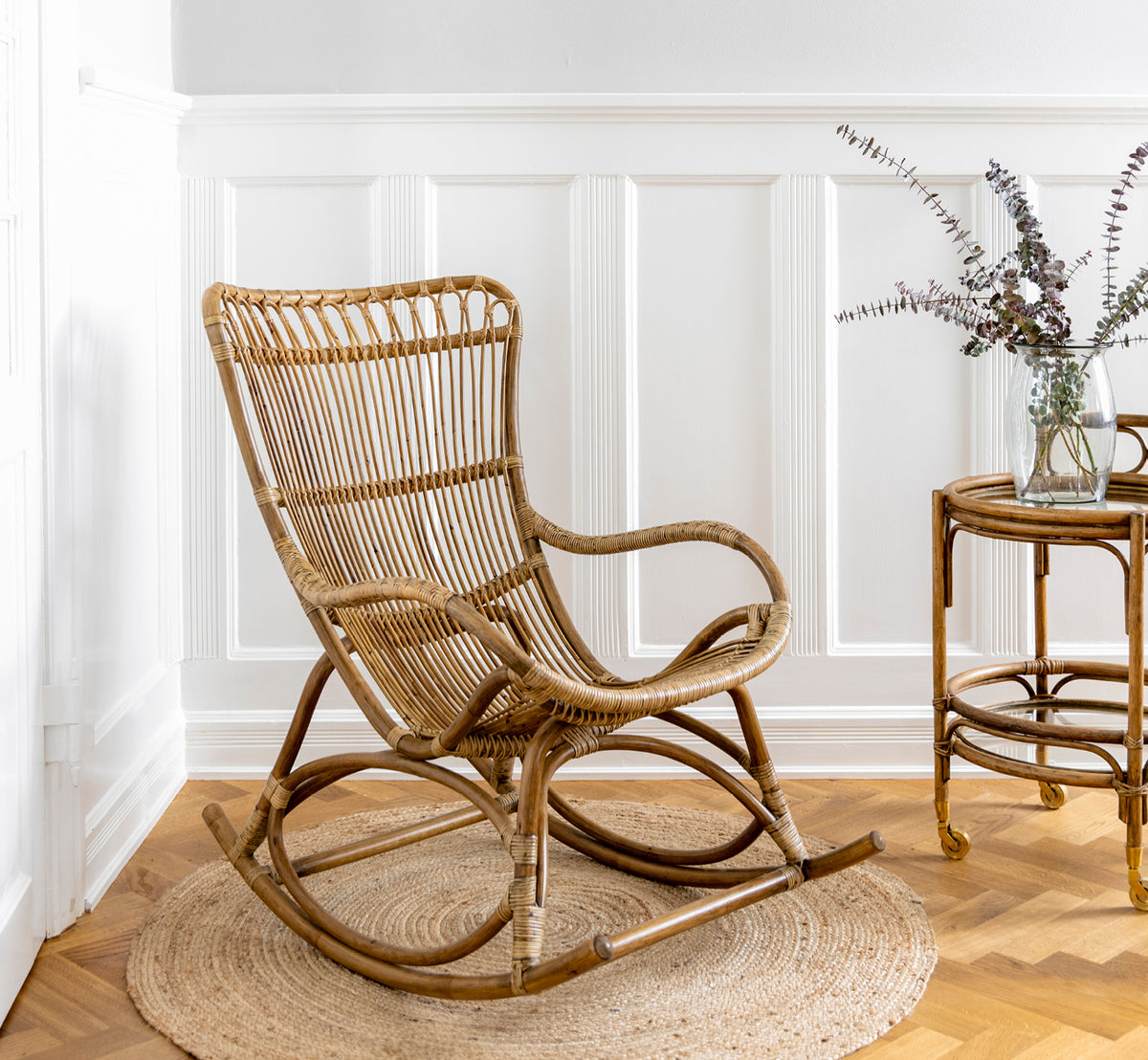 Dette er et livsstilsbillede af Monet Rocking Chair i antique rattan. I et rum med sommerfugletapet og et rattansidebord med glasplade.