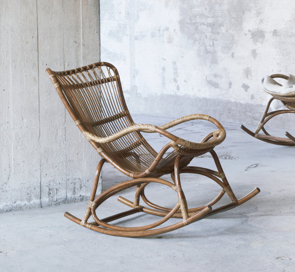 Dette er et livsstilsbillede af Monet Rocking Chair i antique rattan. I et rustikt betonrum med en anden gyngestol i baggrunden.
