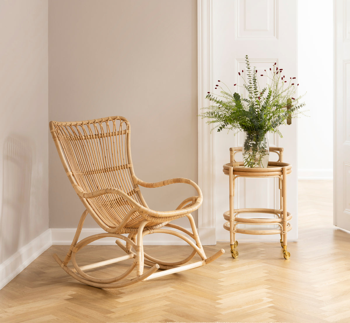Dette er et livsstilsbillede af Monet Rocking Chair i natural rattan. I et hyggeligt hjørne med sildebensgulv og rattanrullebord.