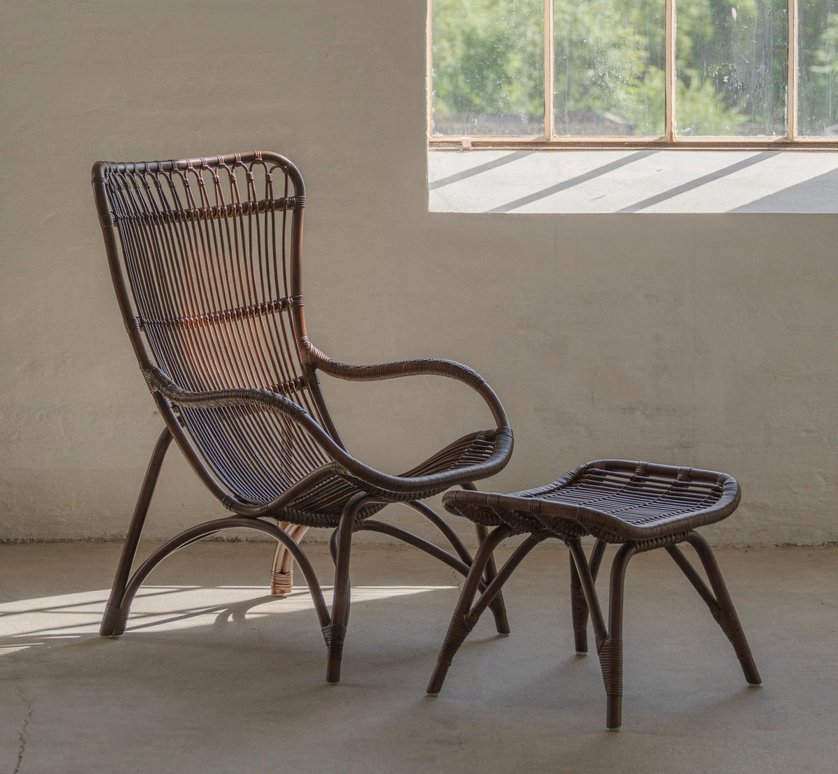 Dette er et livsstilsbillede af Monet Lænestol i brown rattan. Med fodskammel i et solbeskinnet rum med betongulv.