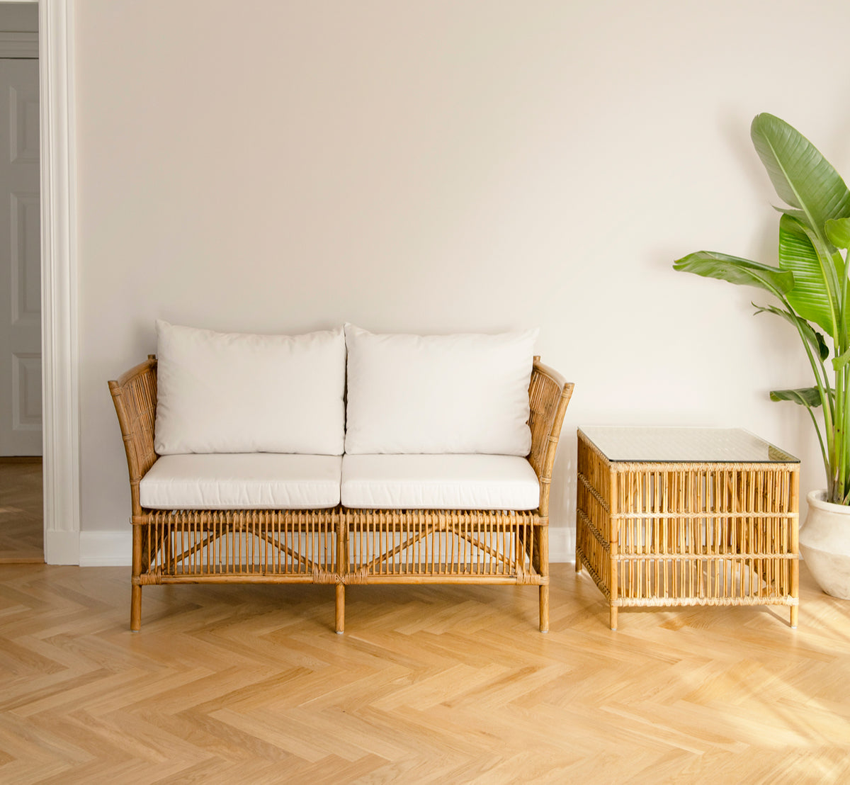 Dette er et livsstilsbillede af Donatello 2-personers sofa i antique rattan, i et minimalistisk lyst interiør med sildebensgulv og en tropisk plante.