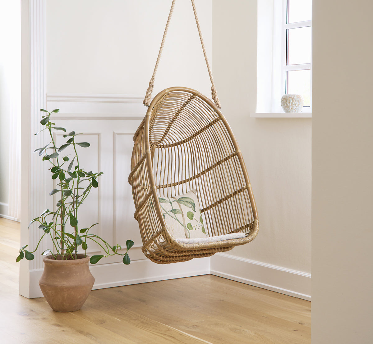Dette er et livsstilsbillede af Renoir Hængestol i antique rattan. Hængende i et lyst panelrum ved siden af en potteplante.