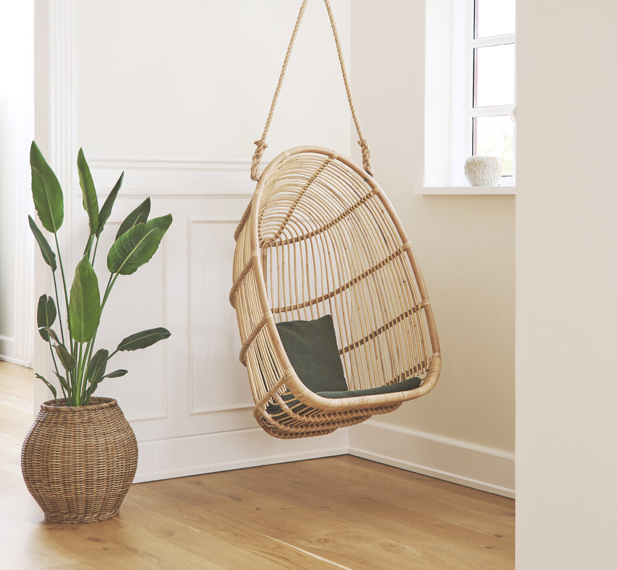 Dette er et livsstilsbillede af Renoir Hængestol i natural rattan. Hængende i en lys panelentré ved siden af en potteplante.