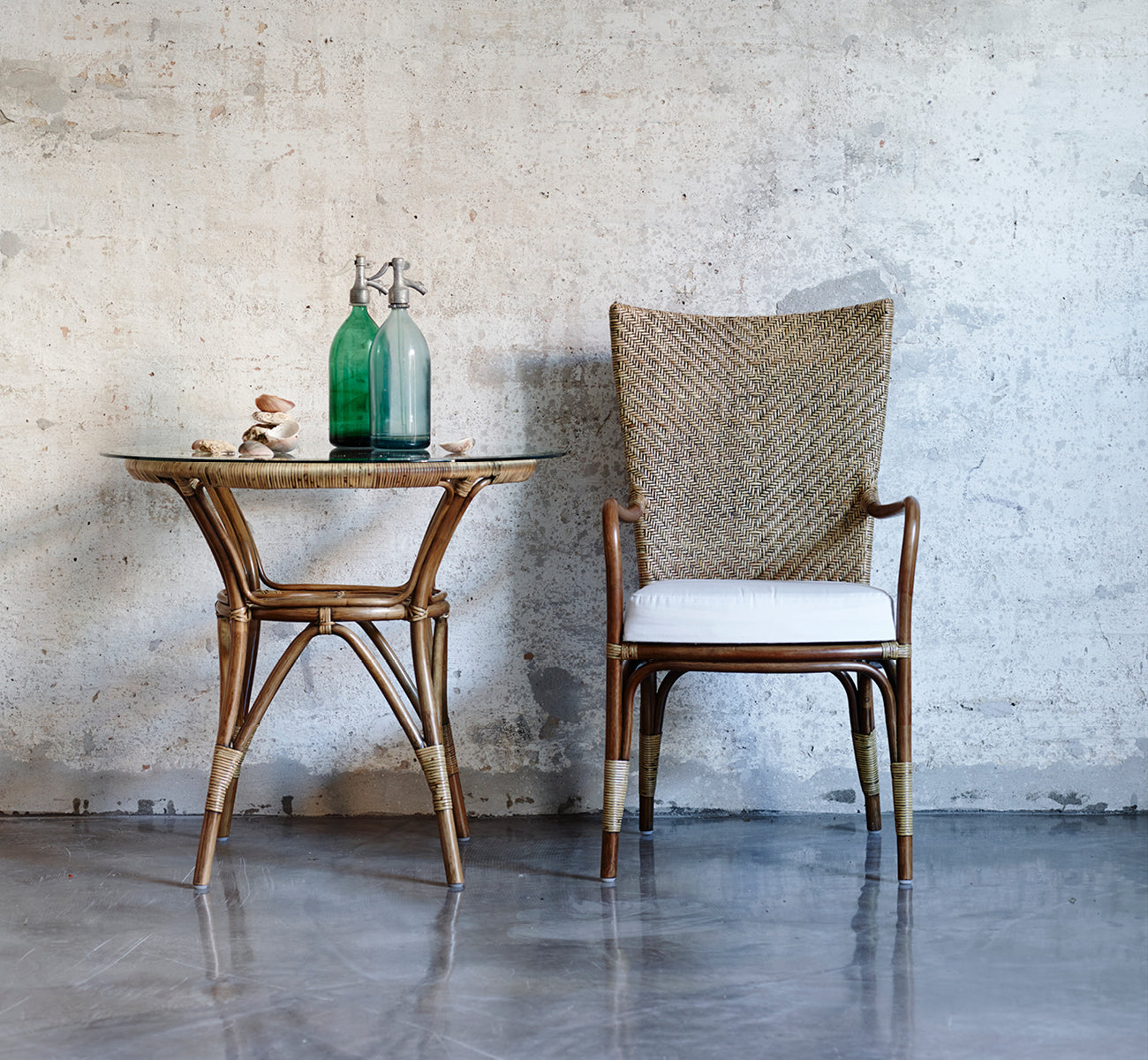 Dette er et livsstilsbillede af Tony Café Table i antique rattan. Kombineret med en højrygget stol mod en rå betonvæg.