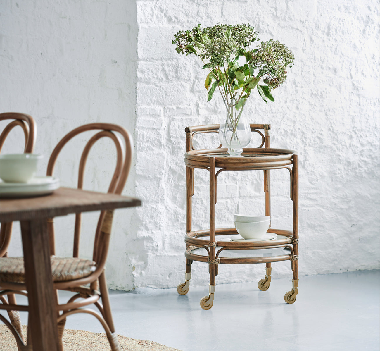 Dette er et livsstilsbillede af Romeo Rullebord i antique rattan. Stylet med hortensiaer og keramik mod en hvid murstensvæg.