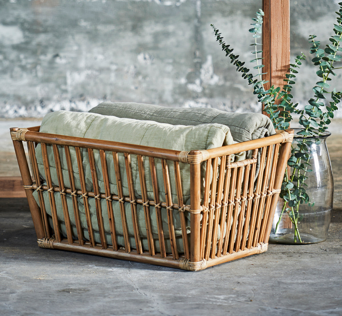 Dette er et livsstilsbillede af Marche Kurv i antique rattan. Stylet med linnedtæpper og eukalyptus i et rum med betonvæg.