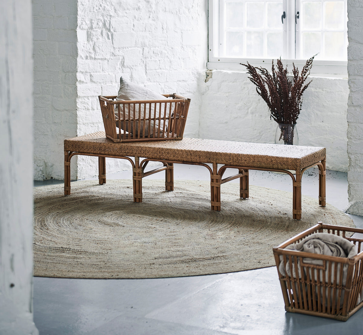 Dette er et livsstilsbillede af Marche Kurv i antique rattan. Placeret på en bænk med puder i et hvidt murstensinteriør med tørrede græsser.