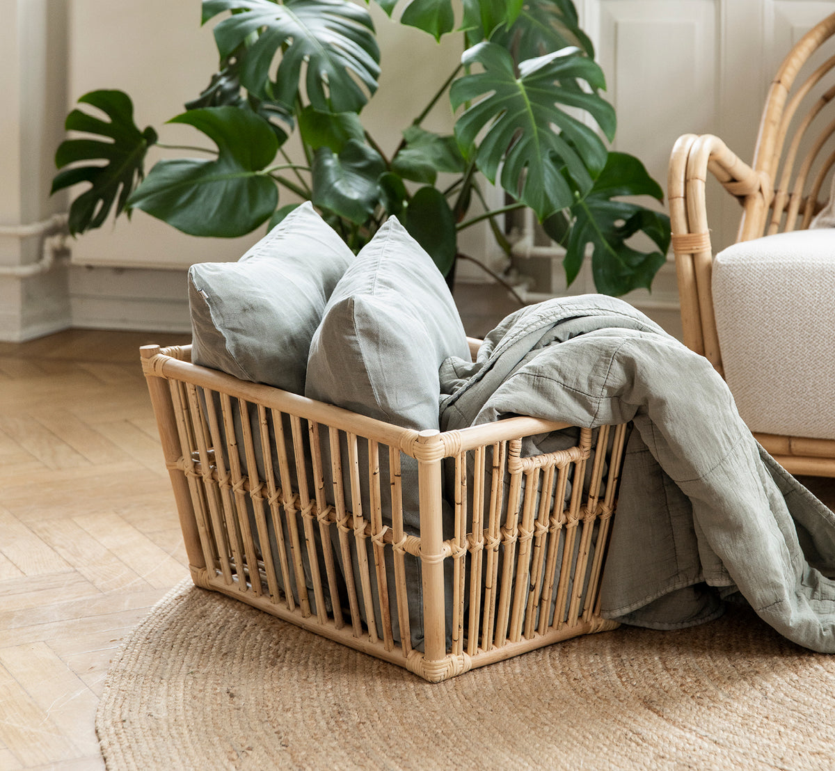 Dette er et livsstilsbillede af Marche Kurv i natural rattan. Fyldt med linnedpuder ved en monsteraplante på egegulv.