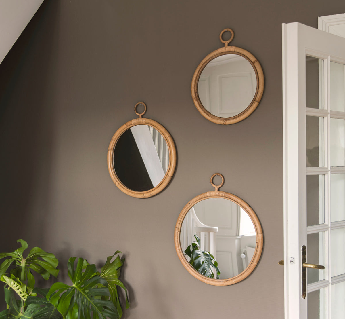 Dette er et livsstilsbillede af Ella Spejl Ø40cm i natural skin on rattan, på en taupe væg i en entré med en monstera og en hvid dør.