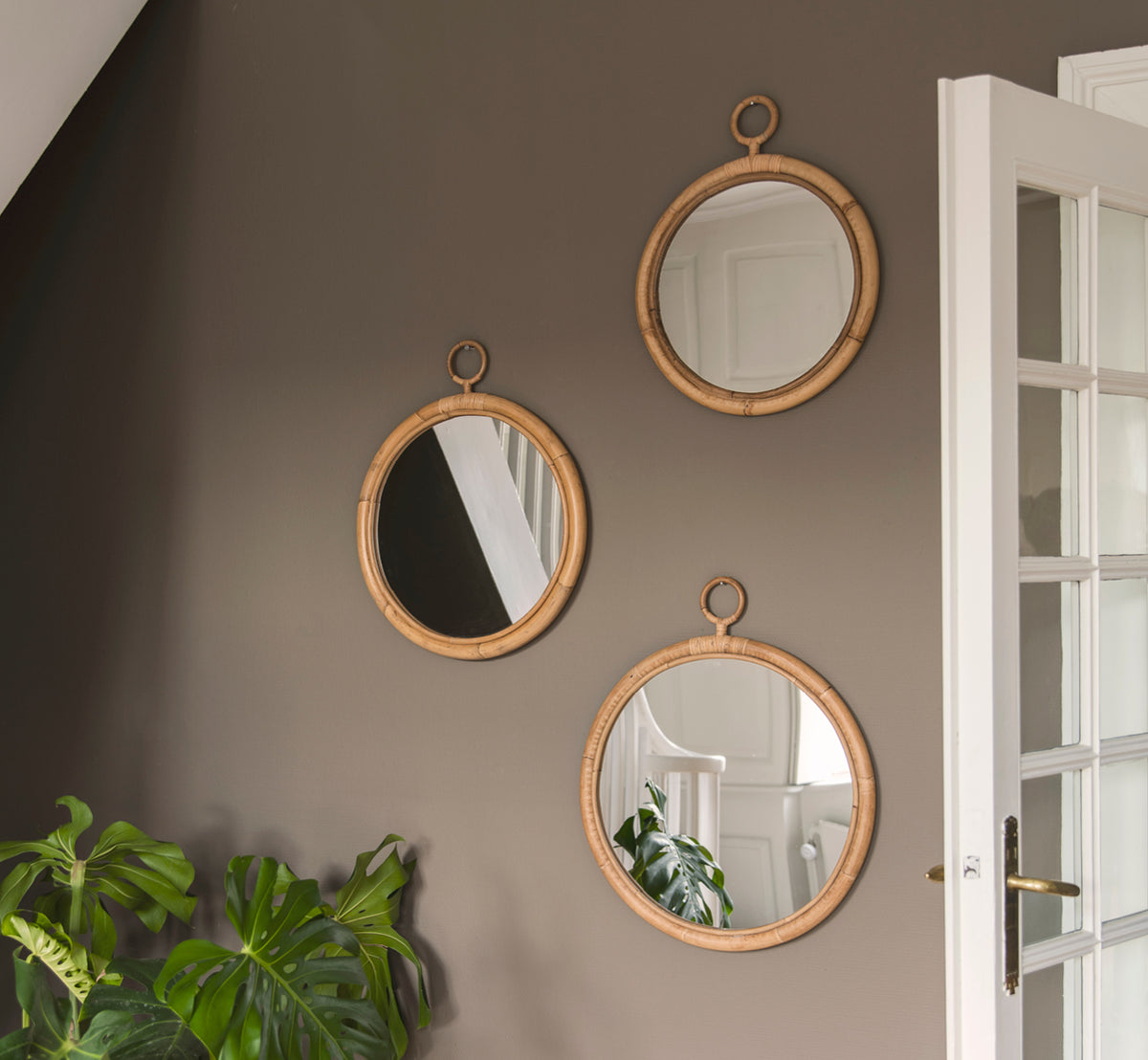 Dette er et livsstilsbillede af Ella Spejl Ø45cm i natural skin on rattan, på en taupe væg i en entré med en monstera og en hvid dør.