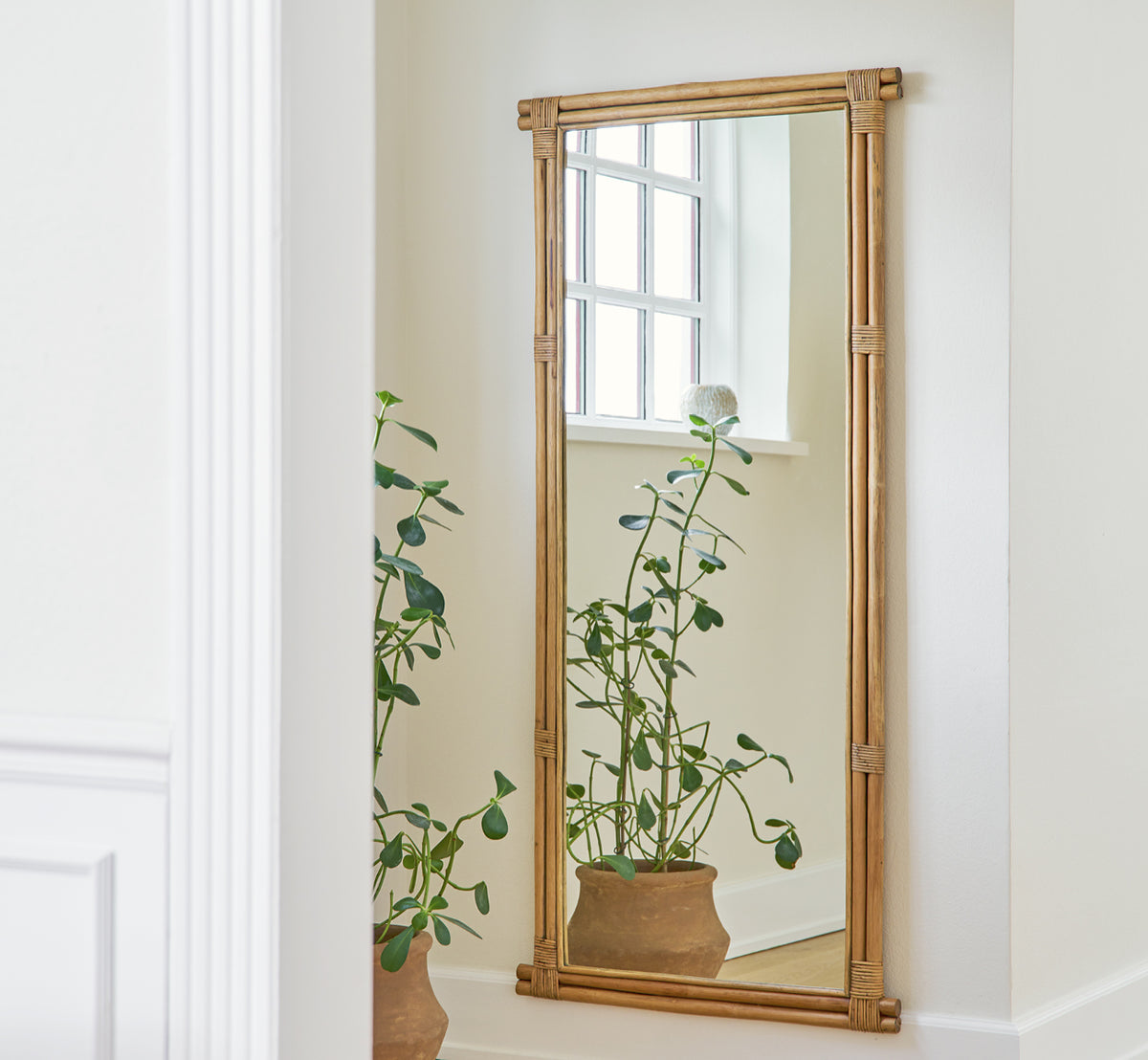 Dette er et livsstilsbillede af Rasmus Mirror 172 cm i antique rattan. Monteret på en hvid væg over en potteplante i en lys korridor.