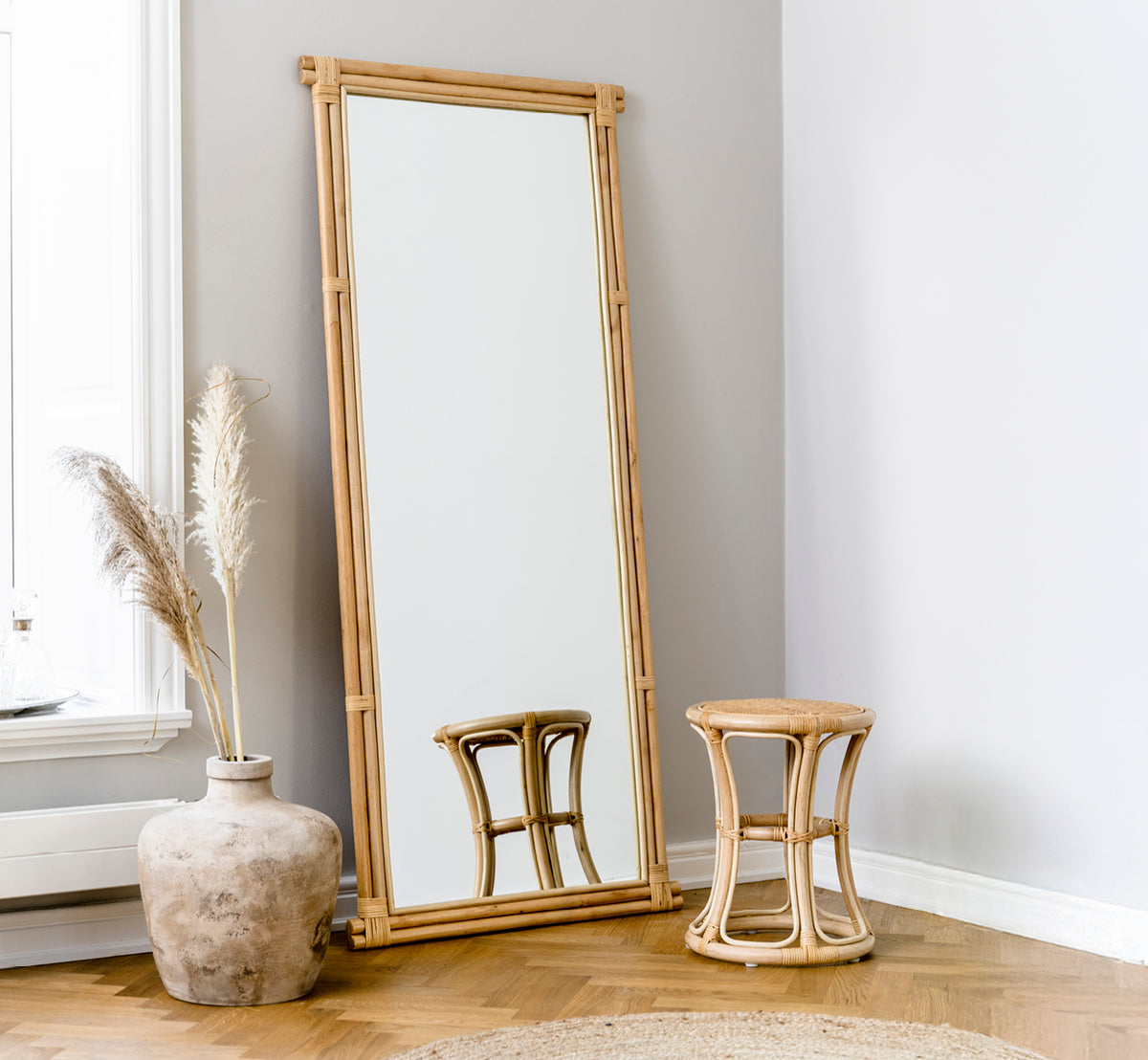 Dette er et livsstilsbillede af Rasmus Mirror 172 cm i natural rattan. Lænet op ad en grå væg ved siden af en skammel og tørret pampasgræs.