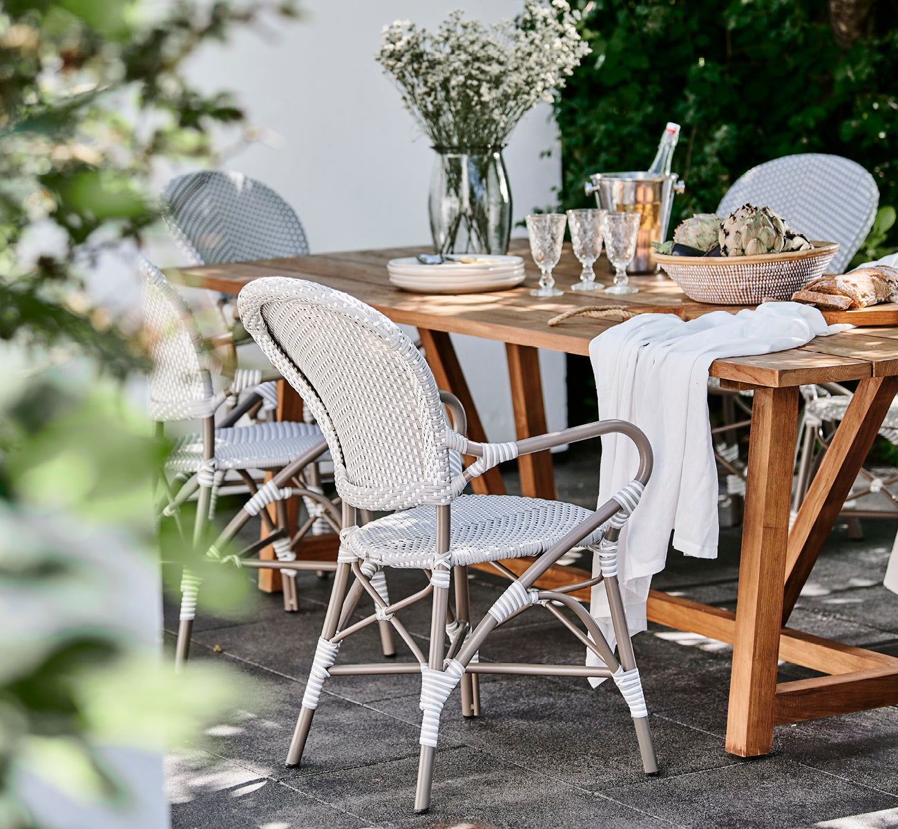 Dette er et livsstilsbillede af Isabell i taupe aluminium frame Grey w white dot ArtFibre, på en frodig haveterrasse med stengulv og en grønklædt.