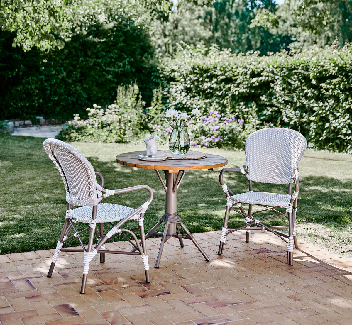Dette er et livsstilsbillede af Isabell i taupe aluminium frame Grey w white dot ArtFibre, på en frodig haveterrasse med stengulv og en grønklædt.