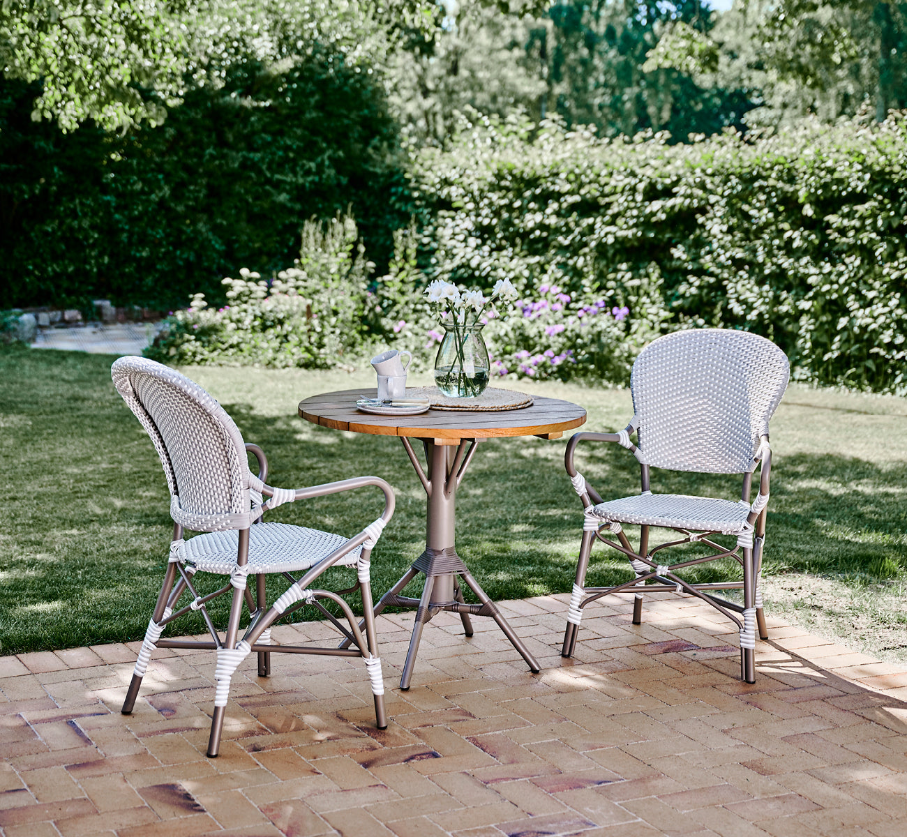 Dette er et livsstilsbillede af Isabell i taupe aluminium frame Grey w white dot ArtFibre, på en frodig haveterrasse med stengulv og en grønklædt.