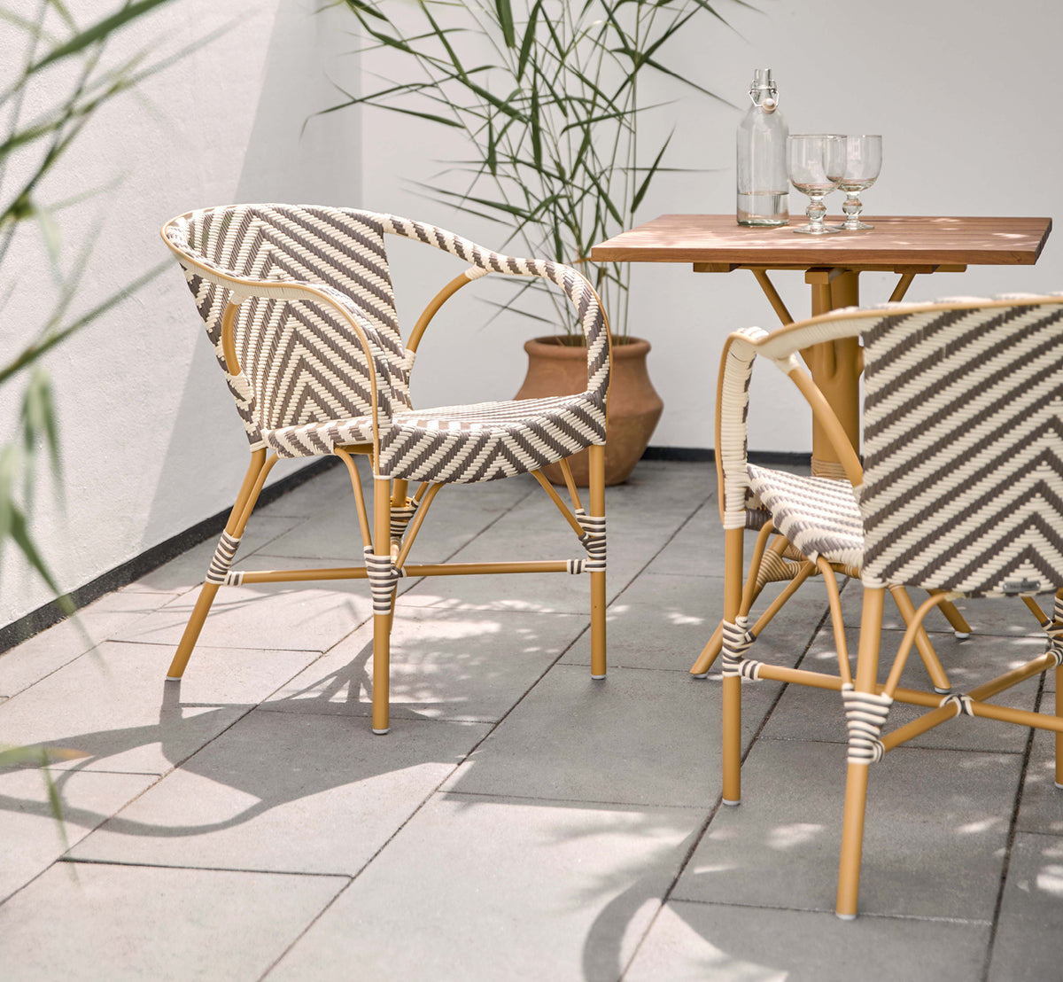 Dette er et livsstilsbillede af Madeleine Udendørs spisebordsstol i almond aluminum frame chevron cappuccino ivory ArtFibre. Solrig terrasse med teakbord.