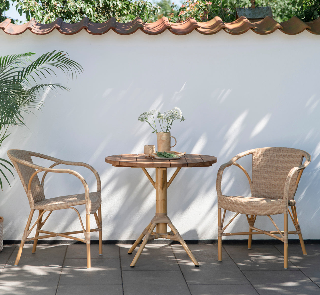Dette er et livsstilsbillede af Madeleine Udendørs spisebordsstol i almond aluminium frame natural ArtFibre. Gårdhave-bistrostemning med rundt teakbord.