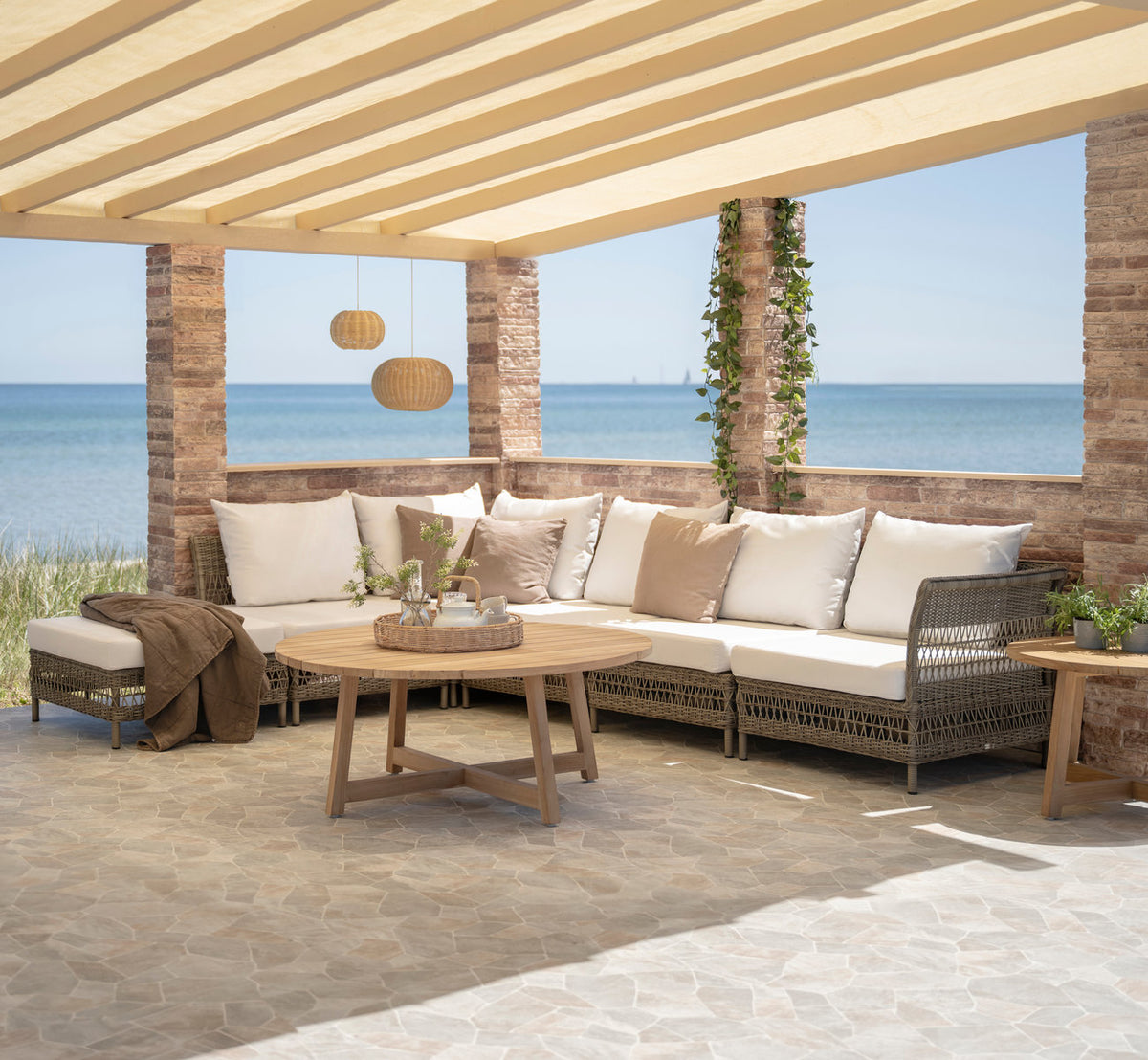Dette er et livsstilsbillede af Mona Udendørs modulsofa-Corner Module i antique grey artfibre aluminium. Under en pergola ved havet.