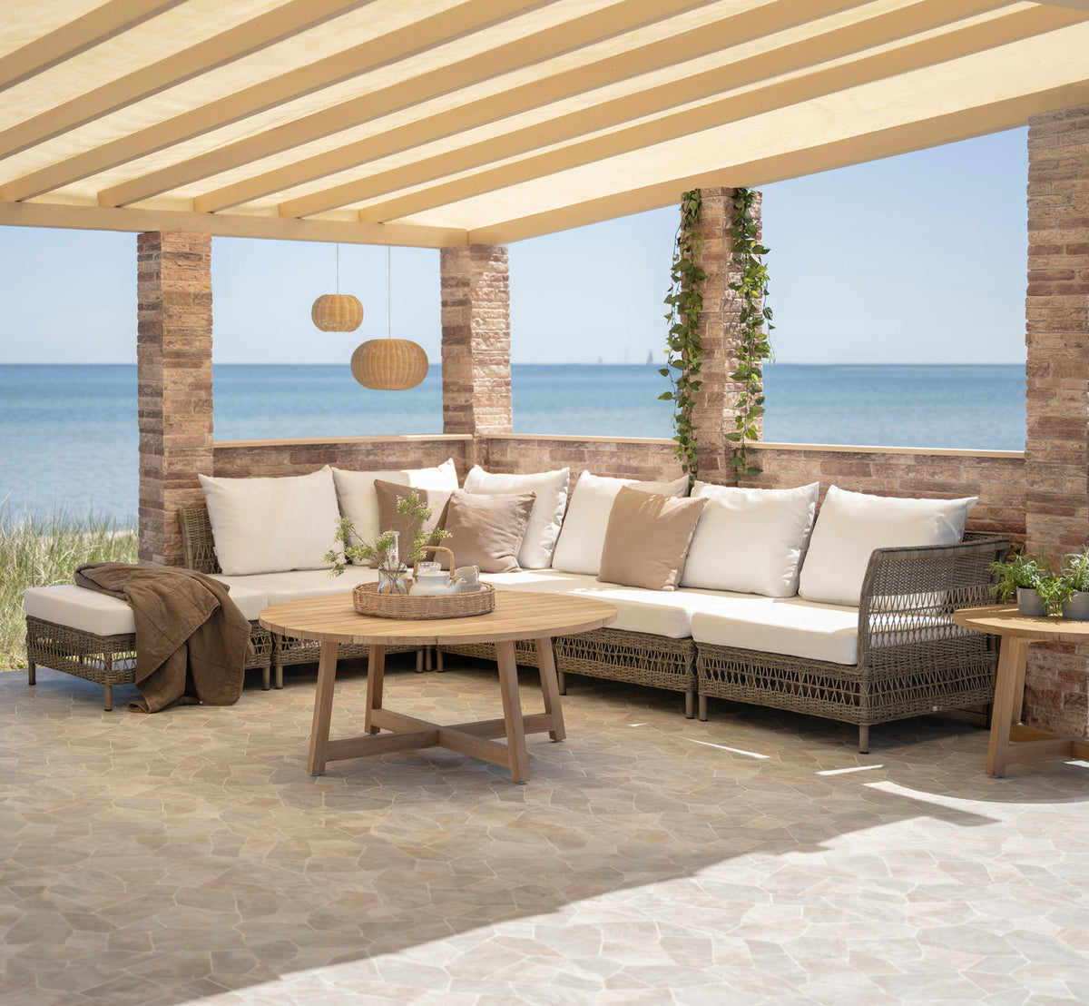 Dette er et livsstilsbillede af Mona Udendørs modulsofa-Pouf Module i antique grey artfibre aluminium. Under en pergola ved havet.