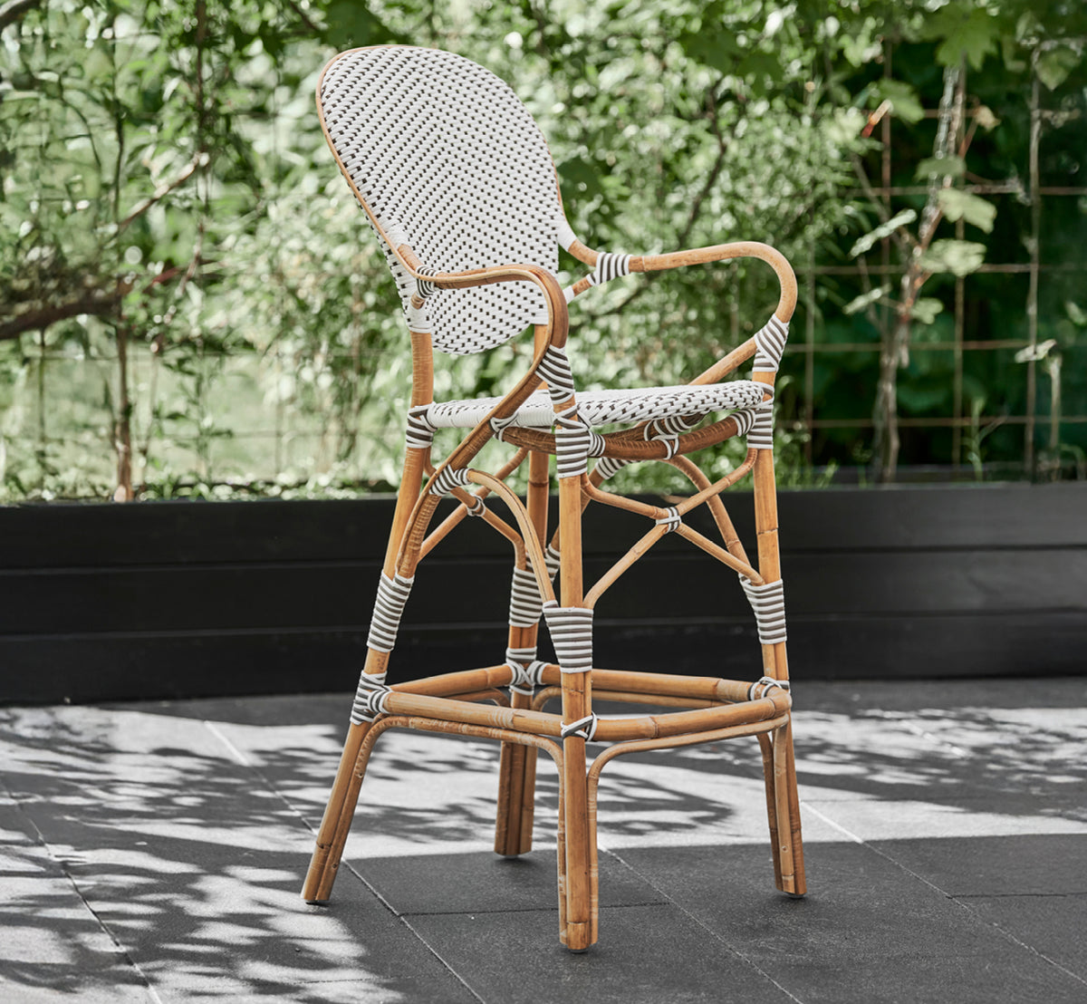 Dette er et livsstilsbillede af Isabell Barstol i white w cappuccino dot ArtFibre Skin on Rattan frame, udendørs mod en frodig grøn havehæk på mørke.