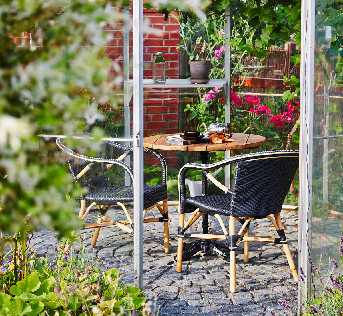 Dette er et livsstilsbillede af Madeleine Spisebordsstol i black ArtFibre skin on rattan frame. Drivhus i haven med brostensgulv og planter.