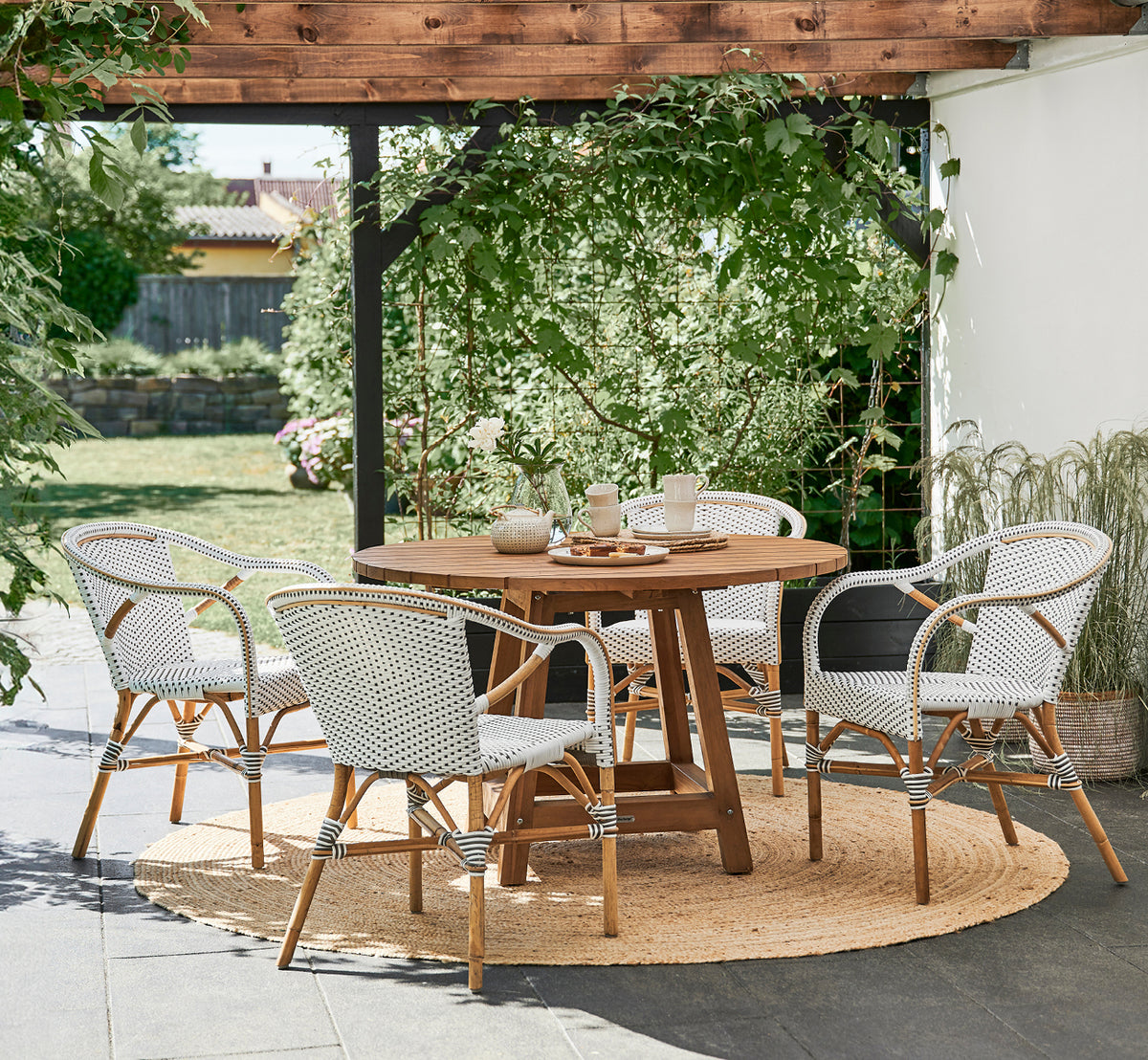 Dette er et livsstilsbillede af Madeleine Spisebordsstol i white w cappuccino dot ArtFibre skin on rattan frame. Havepergola med jutetæppe.