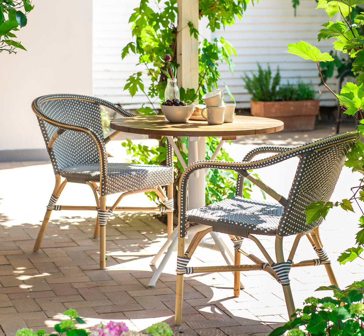 Dette er et livsstilsbillede af Madeleine Spisebordsstol i cappuccino w white dot rattan frame. Skyggefuld haveterrasse med rundt teakbord.