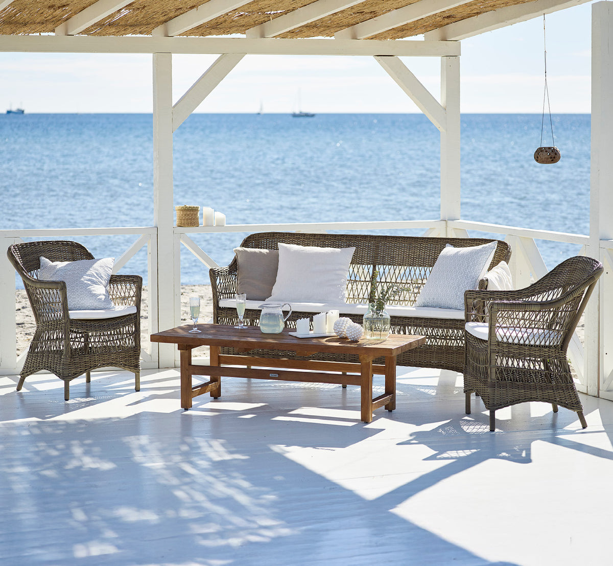 Dette er et livsstilsbillede af Charlot Udendørs stol i antique Grey ArtFibre Aluminium, på en soldrenkt kystterrasse med havudsigt og en hvid.