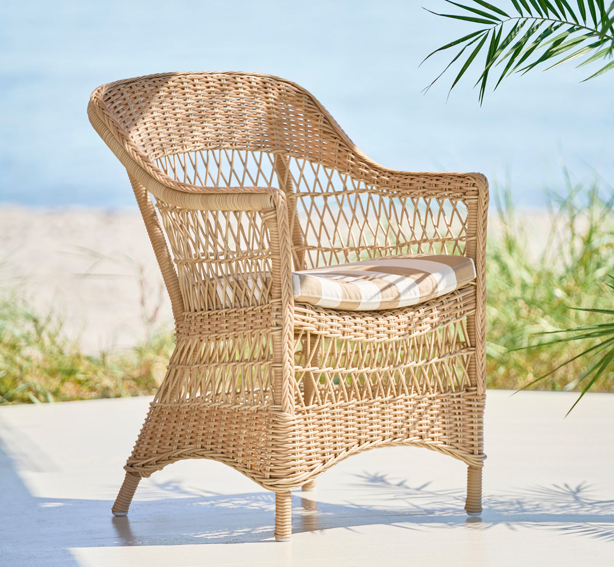 Dette er et livsstilsbillede af Charlot Stol i natural ArtFibre Aluminium, på en soldrenkt kystterrasse med havudsigt og en hvid pergola.