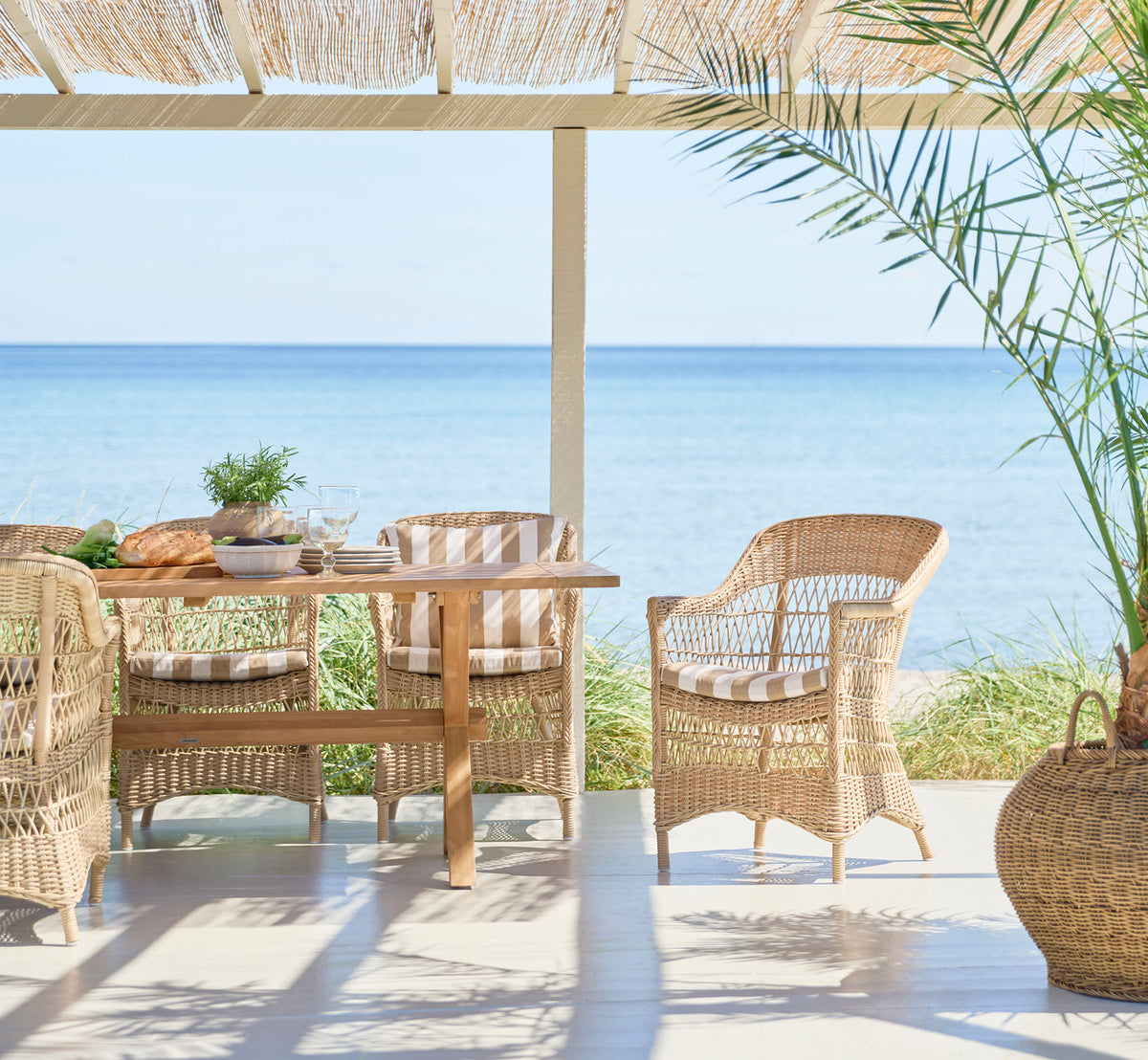 Dette er et livsstilsbillede af Charlot Stol i natural ArtFibre Aluminium, på en soldrenkt kystterrasse med havudsigt og en hvid pergola.