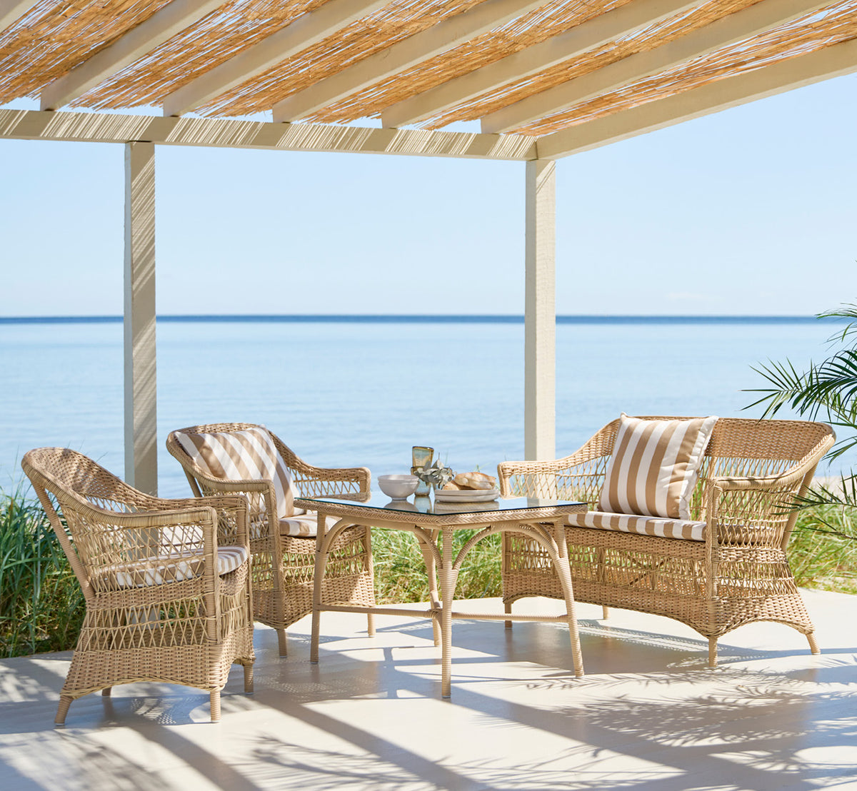 Dette er et livsstilsbillede af Charlot Stol i natural ArtFibre Aluminium, på en soldrenkt kystterrasse med havudsigt og en hvid pergola.
