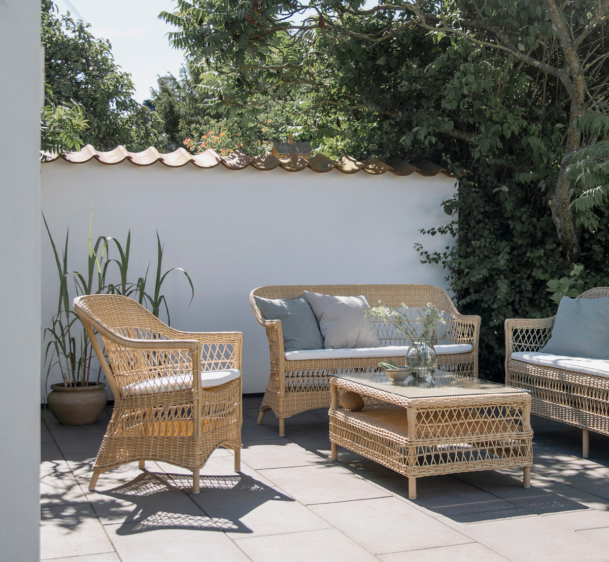 Dette er et livsstilsbillede af Charlot Stol i natural ArtFibre Aluminium, på en soldrenkt kystterrasse med havudsigt og en hvid pergola.