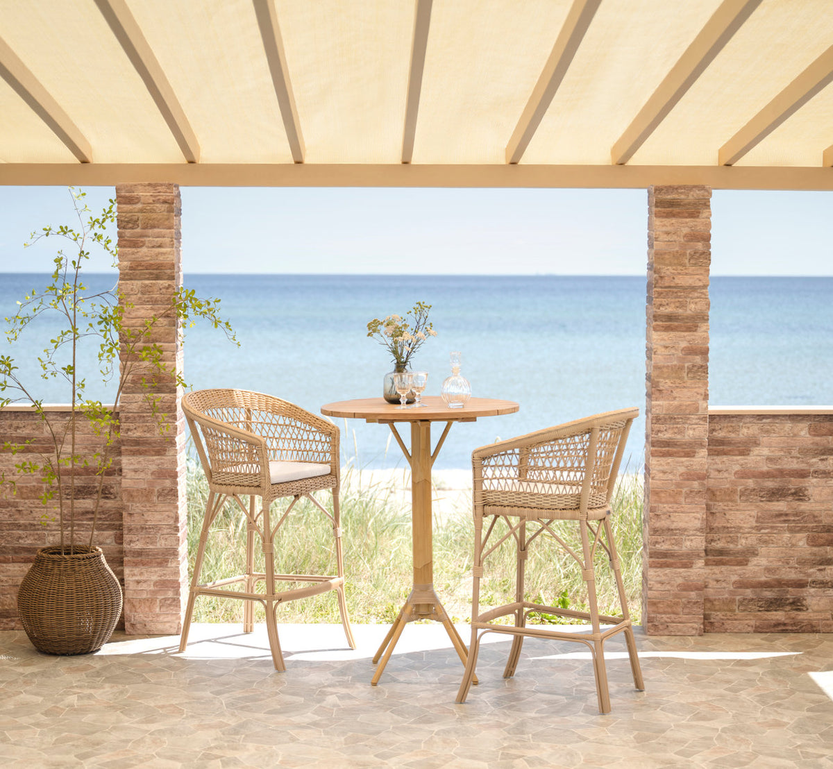 Dette er et livsstilsbillede af Emma Udendørs barstol i natural artfibre aluminium. Baropstilling på en terrasse ved havet.