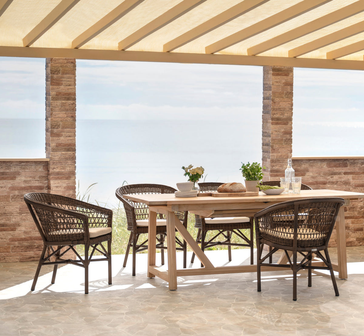 Dette er et livsstilsbillede af Emma Udendørs stol i brown artfibre aluminum. Dækket til frokost på en terrasse ved havet.