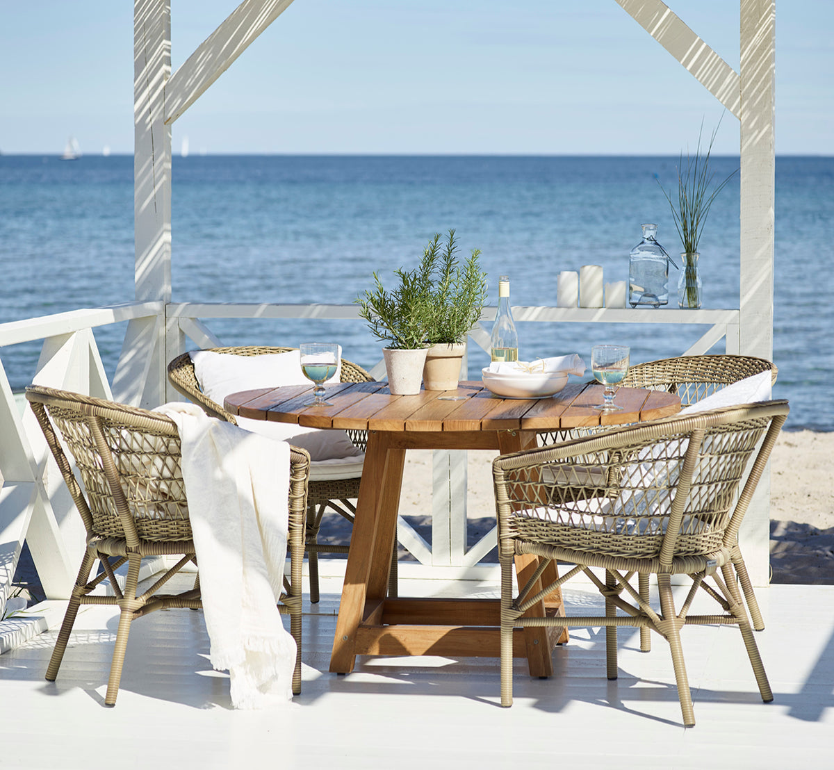 Dette er et livsstilsbillede af Emma Udendørs Spisebordsstol i antique Grey ArtFibre Aluminium, på en lys kystterrasse med sivtag og havudsigt.