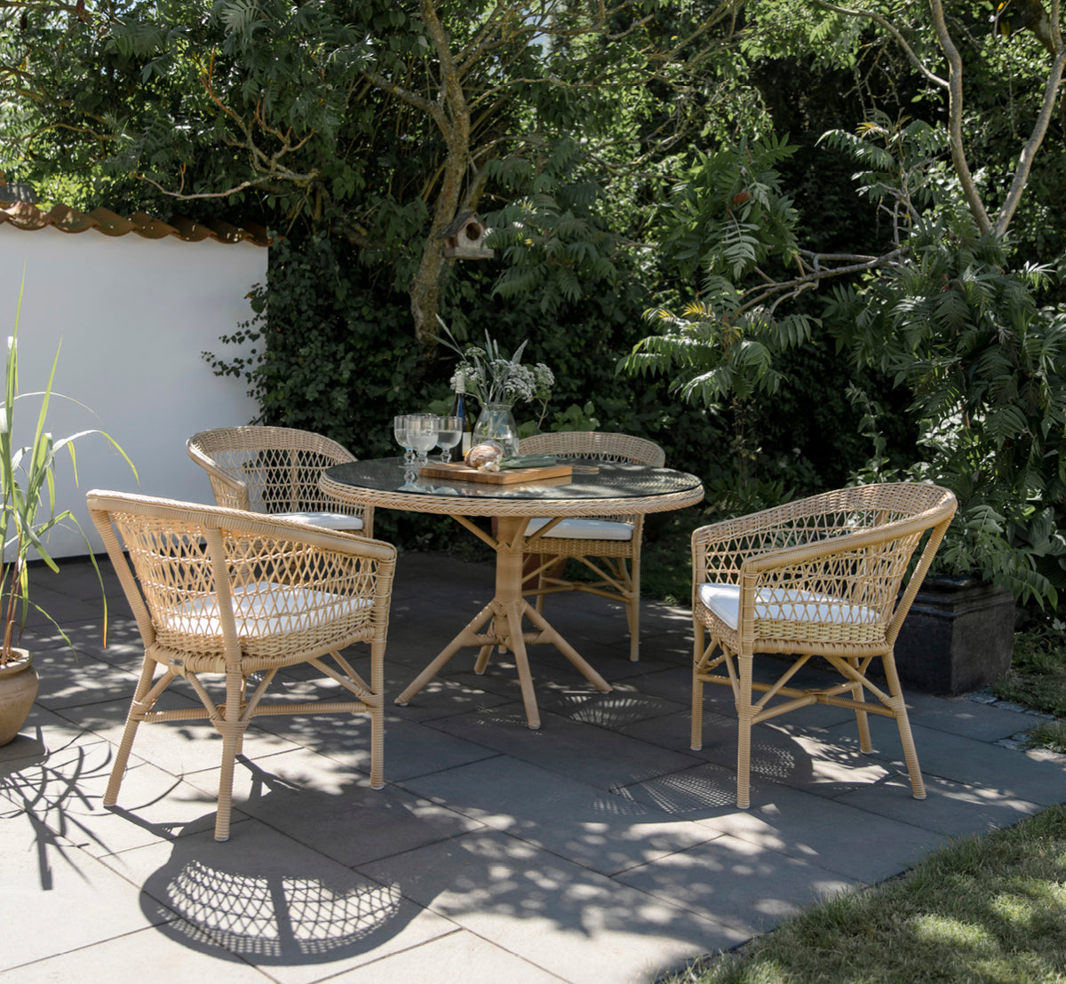 Dette er et livsstilsbillede af Emma Udendørs Spisebordsstol i natural ArtFibre Aluminium, på en lys kystterrasse med sivtag og havudsigt.