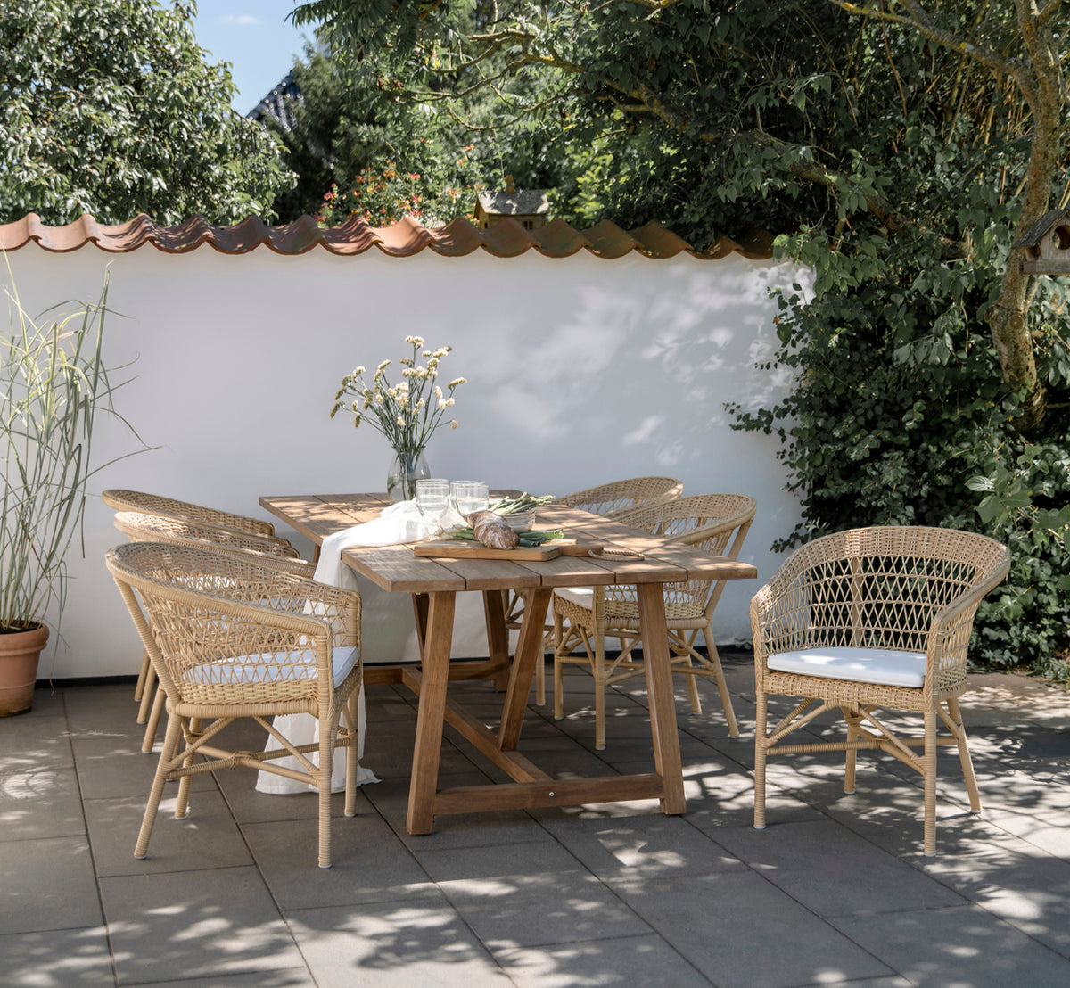 Dette er et livsstilsbillede af Emma Udendørs Spisebordsstol i natural ArtFibre Aluminium, på en lys kystterrasse med sivtag og havudsigt.