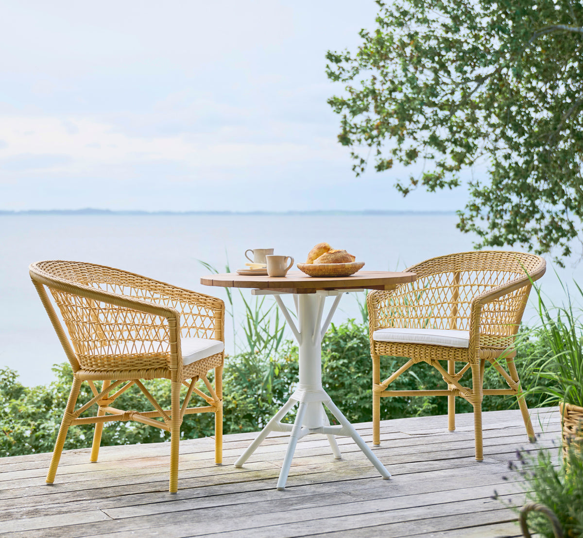 Dette er et livsstilsbillede af Emma Udendørs Spisebordsstol i natural ArtFibre Aluminium, på en lys kystterrasse med sivtag og havudsigt.