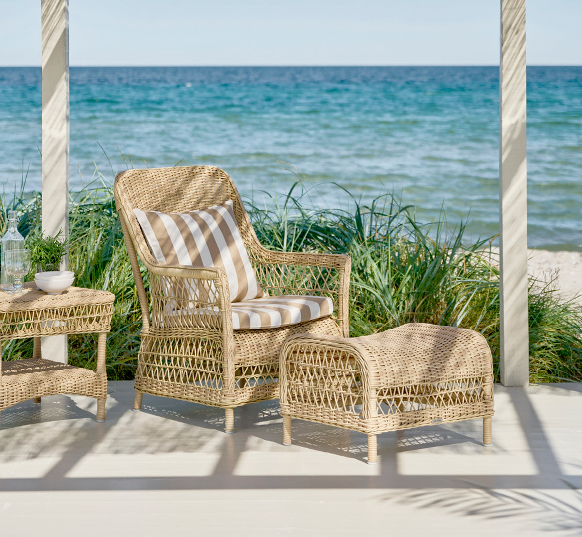 Dette er et livsstilsbillede af Dawn Lænestol i natural ArtFibre Aluminium, på en kystterrasse med havudsigt, strandgræs og hvide søjler.