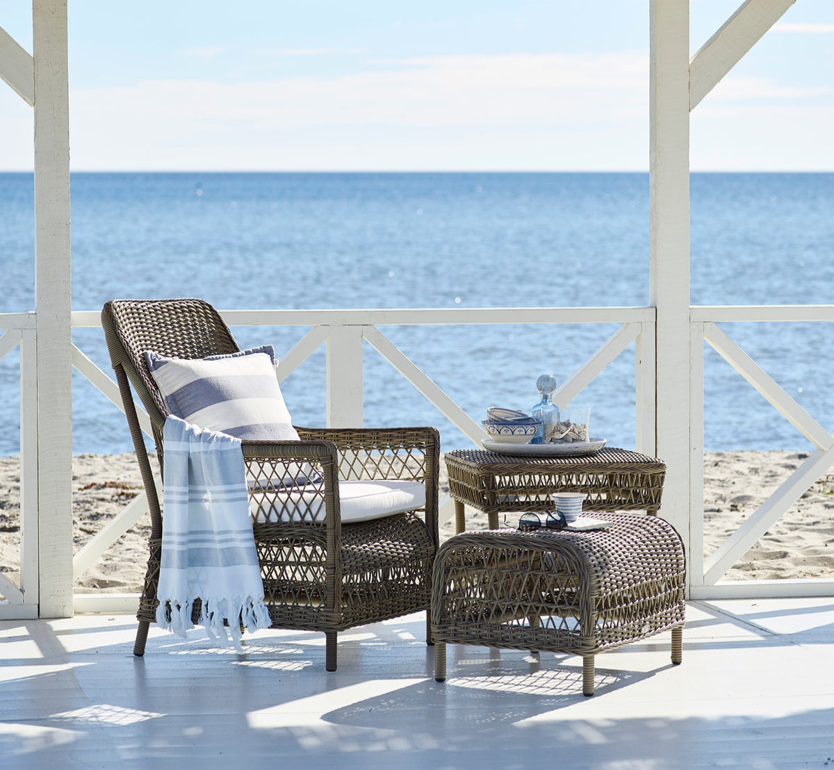Dette er et livsstilsbillede af Dawn Fodskammel i antique Grey ArtFibre Aluminium, på en kystterrasse med havudsigt, strandgræs og hvide søjler.