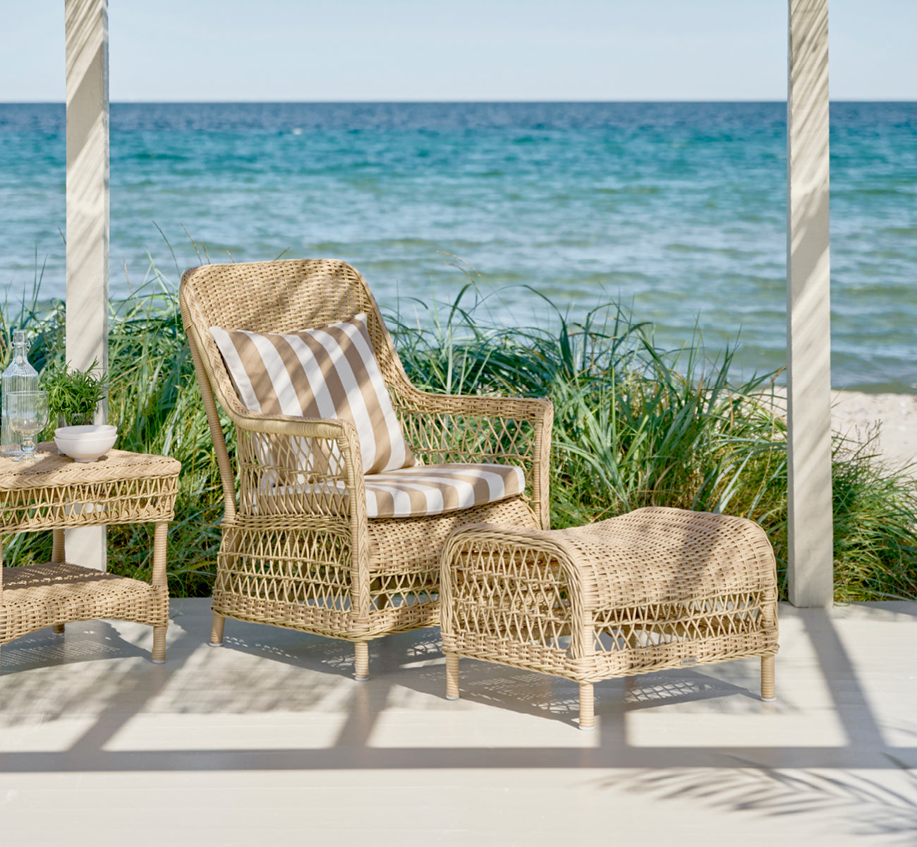 Dette er et livsstilsbillede af Dawn Fodskammel i natural ArtFibre Aluminium, på en kystterrasse med havudsigt, strandgræs og hvide søjler.