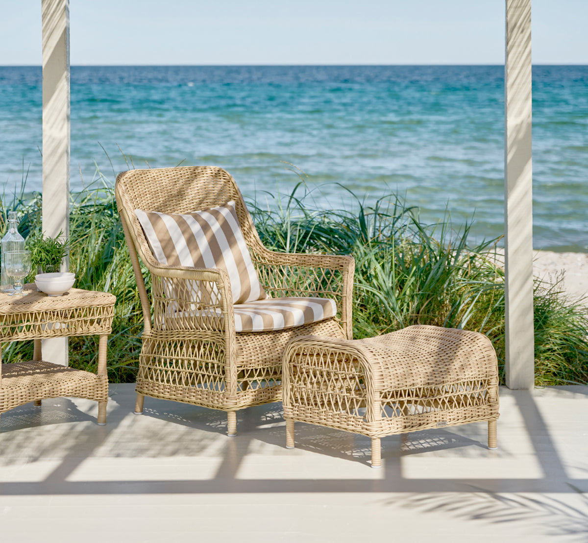 Dette er et livsstilsbillede af Dawn Fodskammel i natural ArtFibre Aluminium, på en kystterrasse med havudsigt, strandgræs og hvide søjler.