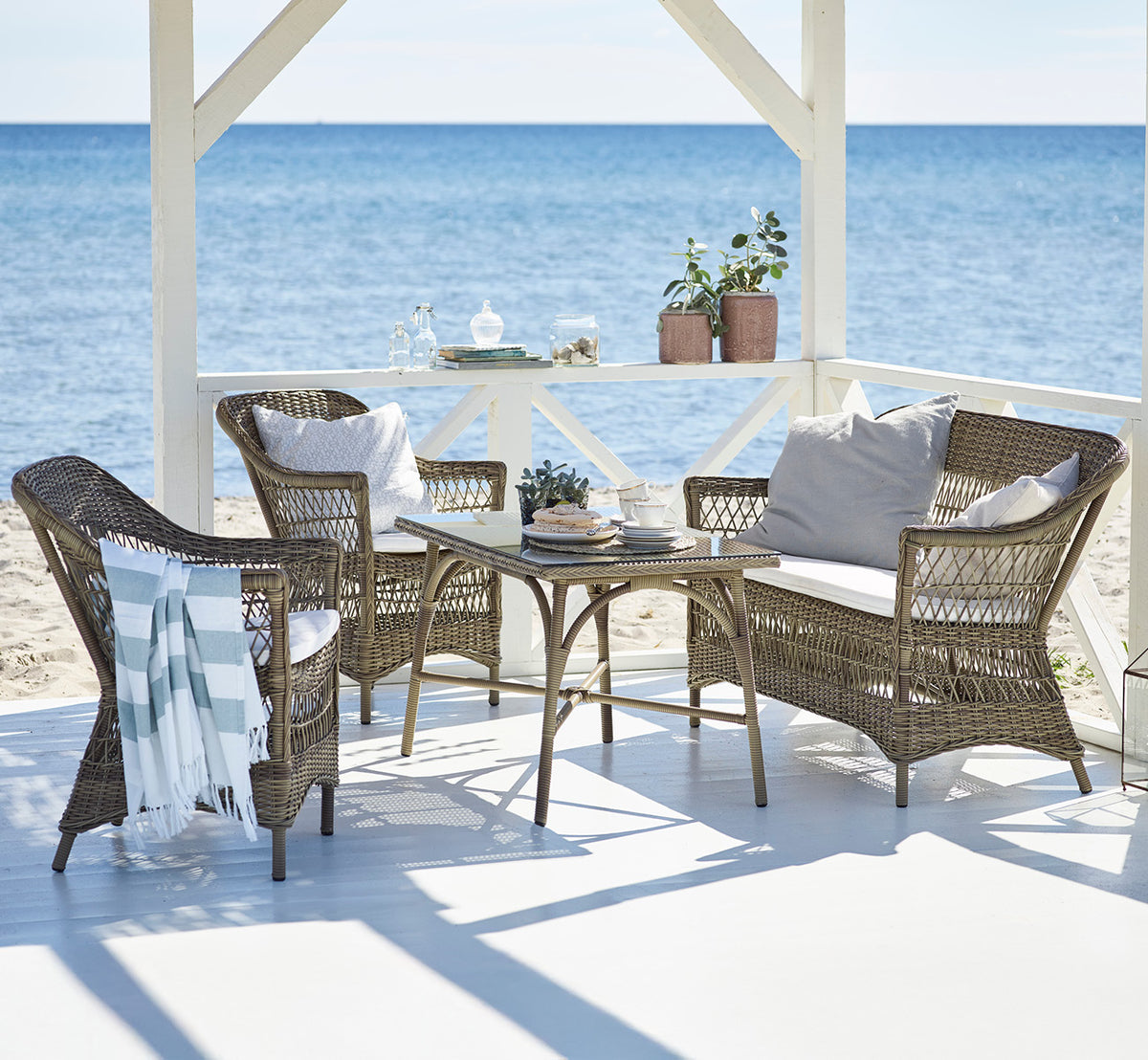 Dette er et livsstilsbillede af Charlot 2-personers sofa i antique Grey ArtFibre Aluminium, på en kystterrasse med sivtag, havudsigt og stribede.