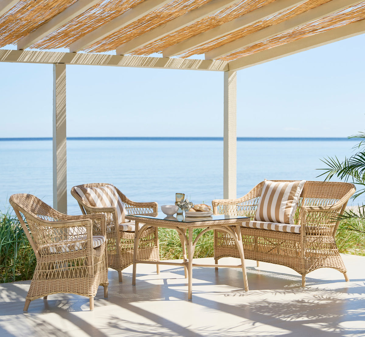 Dette er et livsstilsbillede af Charlot 2-personers sofa i natural ArtFibre Aluminium, på en kystterrasse med sivtag, havudsigt og stribede puder.