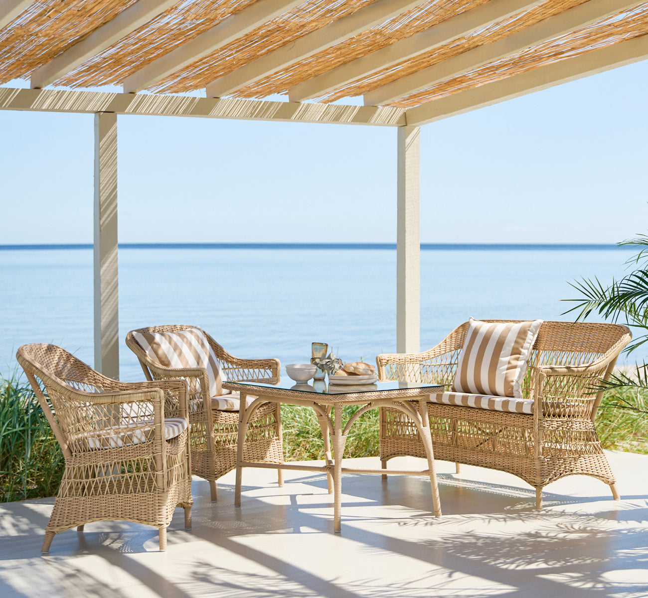 Dette er et livsstilsbillede af Charlot 2-personers sofa i natural ArtFibre Aluminium, på en kystterrasse med sivtag, havudsigt og stribede puder.